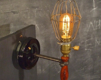 May include: A vintage-style wall lamp with a caged light bulb and a wooden handle labeled "Forester". The lamp is mounted on a black metal plate and has a black wire cord.