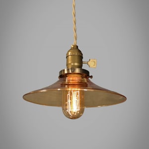 Puede incluir: Lámpara colgante de estilo vintage con una pantalla de cristal color ámbar y una estructura de latón. La bombilla es visible, emitiendo un brillo suave. El cable es trenzado y de color crema.