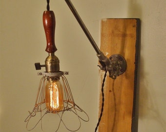 May include: A vintage-style wall-mounted light fixture with a wooden handle, a metal cage shade, and a  light bulb. The fixture is attached to a wooden board with a metal clamp.