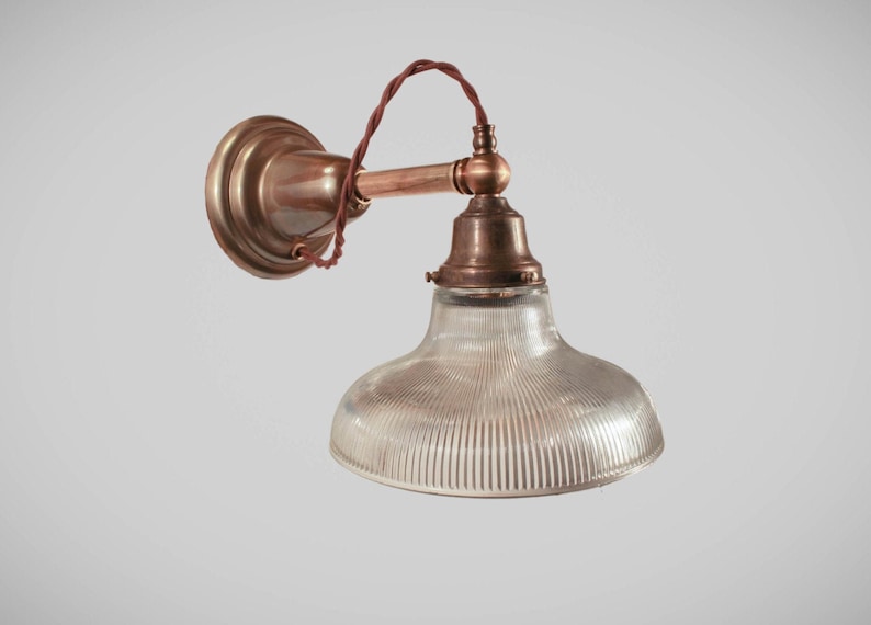 May include: Vintage-style wall sconce with a clear, ribbed glass shade and a copper-toned metal fixture. The fixture includes a round wall mount, a curved arm, and a decorative bulb holder. The wiring is a twisted brown cord.
