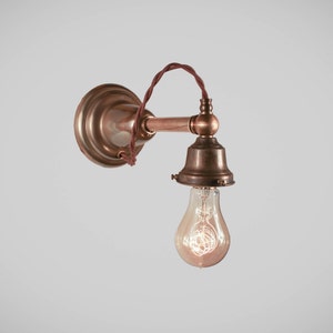May include: Vintage-style wall sconce with a copper finish. The fixture features a round backplate, a horizontal arm, and a bell-shaped shade holding a clear Edison bulb. The wiring is a twisted brown cord.