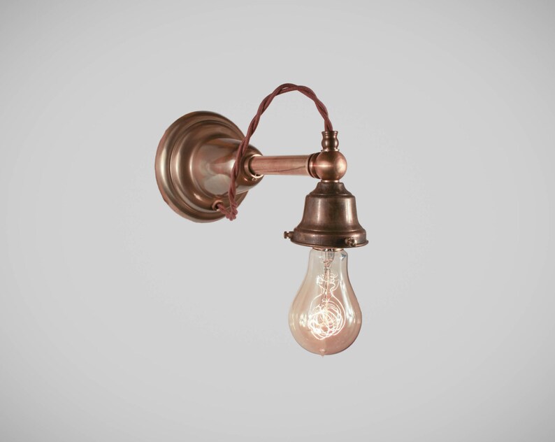 May include: Vintage-style wall sconce with a copper finish. The fixture features a round backplate, a horizontal arm, and a bell-shaped shade holding a clear Edison bulb. The wiring is a twisted brown cord.