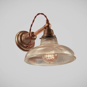 May include: Vintage-style wall sconce with a clear, ribbed glass shade and a warm, exposed filament bulb. The fixture features a brass-toned mounting plate and arm, with a brown twisted cord. The design evokes an industrial or retro aesthetic.