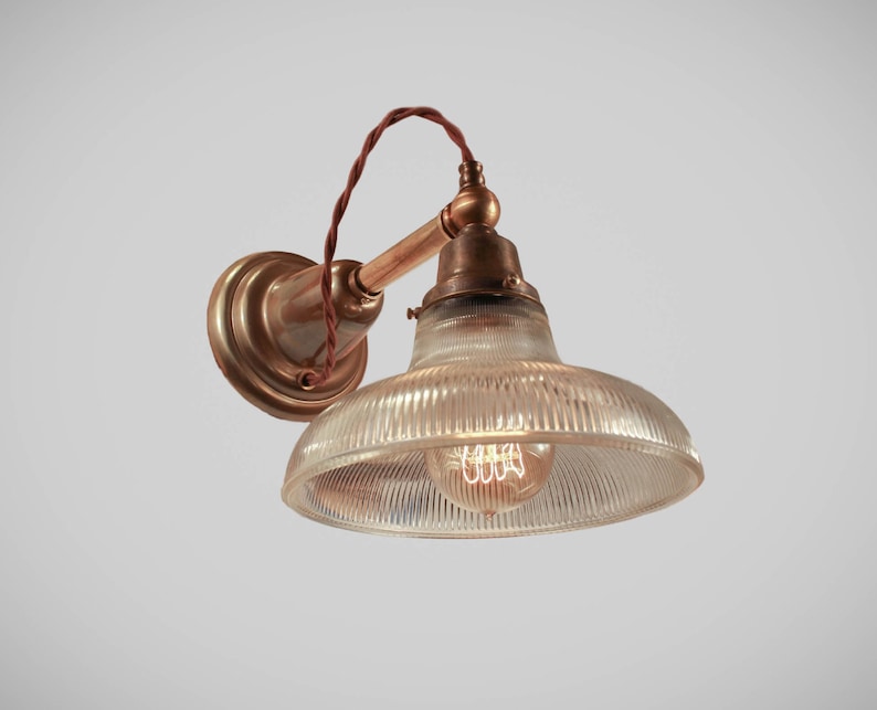 May include: Vintage-style wall sconce with a clear, ribbed glass shade and a warm, exposed filament bulb. The fixture features a brass-toned mounting plate and arm, with a brown twisted cord. The design evokes an industrial or retro aesthetic.