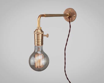Könnte beinhalten: Industrielle Wandleuchte mit Messing-Finish. Die Leuchte hat einen gebogenen Arm, eine runde Wandplatte und eine Edison-Glühbirne im Vintage-Stil. Ein braunes, gedrehtes Elektrokabel hängt herunter.