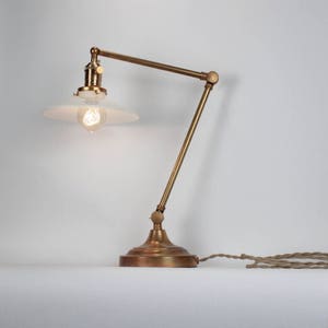 Op de afbeelding: Een messing bureaulamp met een verstelbare scharnierarm en een brede, witte conische kap. De lamp heeft een ronde voet, een fitting in vintage-stijl met een kleine draaischakelaar en een heldere Edison-gloeilamp met een zichtbare gloeidraad.