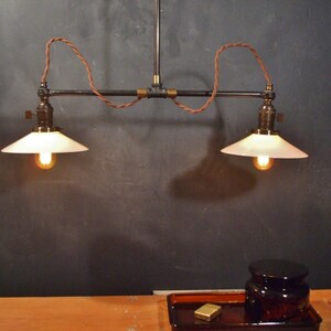 May include: Two white glass lampshades with a black metal pipe fixture and brown wire hanging from a ceiling. The lampshades are lit with warm white light bulbs.