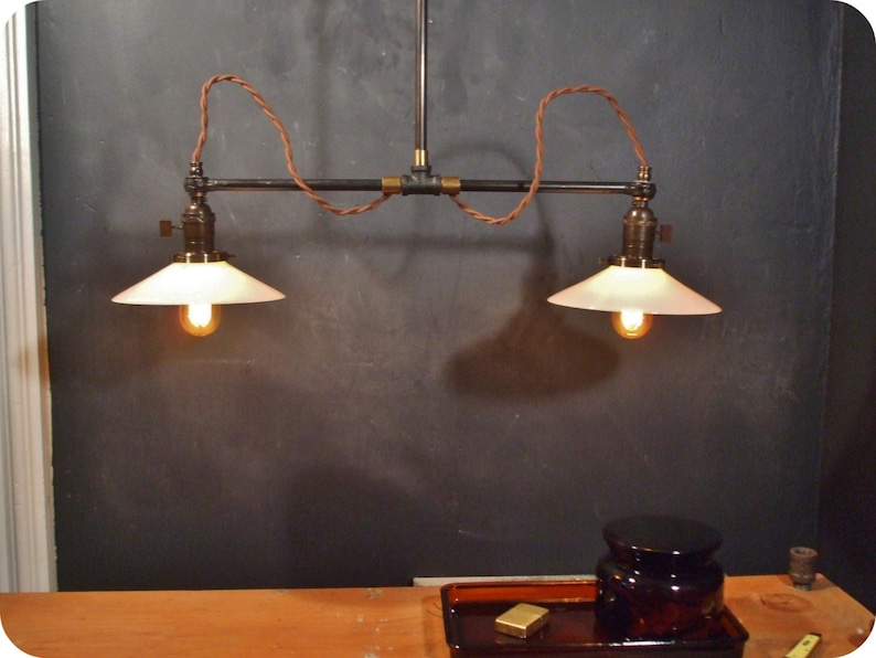 May include: Two white glass lampshades with a black metal pipe fixture and brown wire hanging from a ceiling. The lampshades are lit with warm white light bulbs.