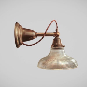 May include: A vintage-style wall-mounted lamp with a brass finish. The lamp features a clear, ribbed glass shade and a brown, twisted electrical cord. The design is reminiscent of industrial or antique lighting fixtures.