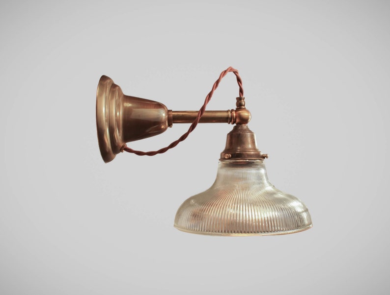 May include: A vintage-style wall-mounted lamp with a brass finish. The lamp features a clear, ribbed glass shade and a brown, twisted electrical cord. The design is reminiscent of industrial or antique lighting fixtures.