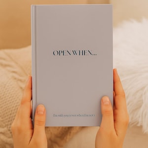 May include: A gray journal with the words "OPEN WHEN..." printed on the cover. Below the title, the text reads "I'm with you (even when I'm not)". The journal is held open by a person.
