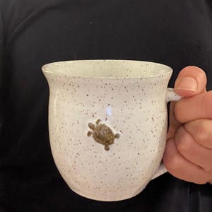May include: White ceramic mug with a speckled finish and a brown turtle design. The mug has a rounded shape with a handle and is held by a person. The mug is a unique piece of pottery.
