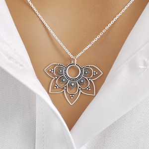 May include: A silver-colored pendant necklace featuring a floral design. The pendant has a detailed, openwork pattern with a central circle and petal-like shapes. The necklace is worn over a white button-down shirt.