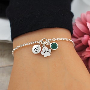 May include: A silver chain bracelet with a charm of a paw print, a green gemstone, and a silver disc with the letter "B" engraved on it.