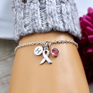 May include: Silver charm bracelet with a pink ribbon, a pink crystal, and a charm with the letter "H".