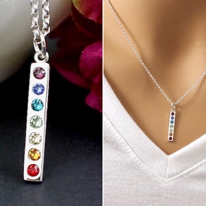 May include: A silver bar pendant necklace featuring seven colorful gemstones. The gemstones are arranged vertically in a row, displaying a spectrum of colors. The necklace is shown on a white background and against a person's white v-neck shirt.