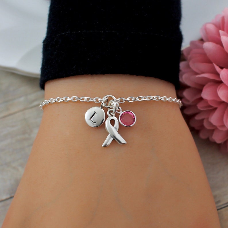 May include: Silver chain bracelet with a pink ribbon charm, a small pink crystal charm, and a silver charm with the letter L.