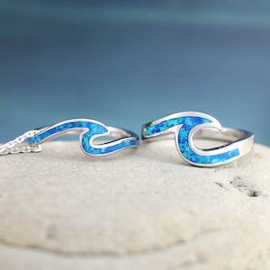 Puede incluir: Un conjunto de anillo y collar de plata con incrustaciones de &oacute;palo azul en forma de ola. El dise&ntilde;o de la ola es un s&iacute;mbolo popular del oc&eacute;ano y la playa.