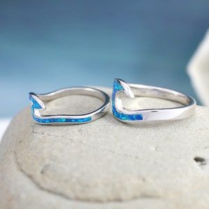 Puede incluir: Dos anillos de plata con una incrustaci&oacute;n de &oacute;palo azul en forma de gancho. Los anillos est&aacute;n sobre una superficie de piedra gris.