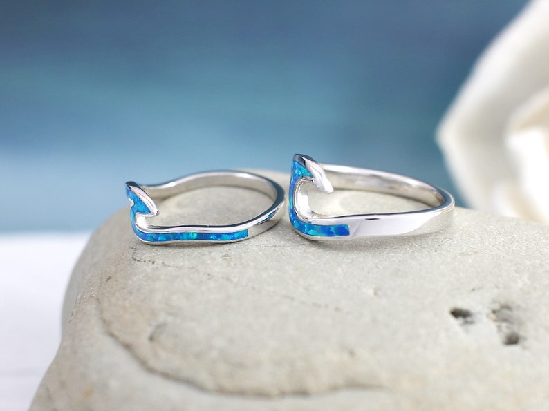 Puede incluir: Dos anillos de plata con una incrustaci&oacute;n de &oacute;palo azul en forma de gancho. Los anillos est&aacute;n sobre una superficie de piedra gris.
