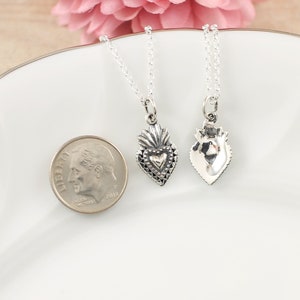 May include: Two silver necklaces with heart-shaped pendants. The pendants are detailed with a textured design and a smaller heart in the center. The necklaces are displayed on a white plate with a silver dime for scale.