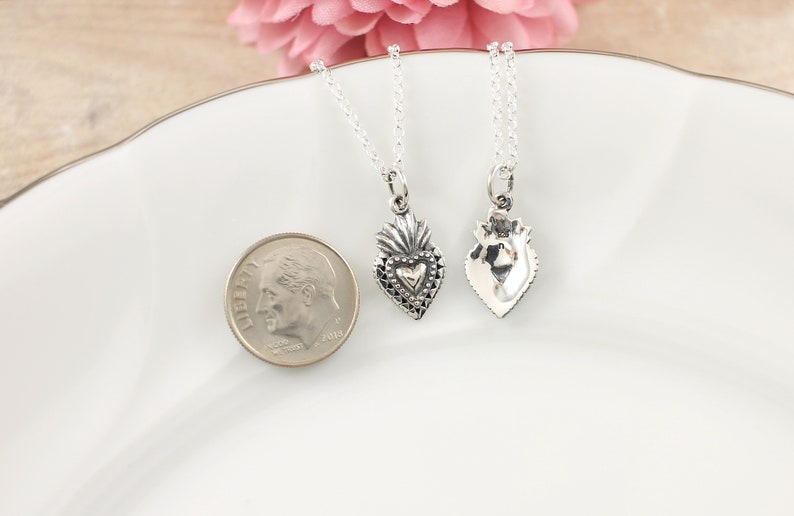 May include: Two silver necklaces with heart-shaped pendants. The pendants are detailed with a textured design and a smaller heart in the center. The necklaces are displayed on a white plate with a silver dime for scale.