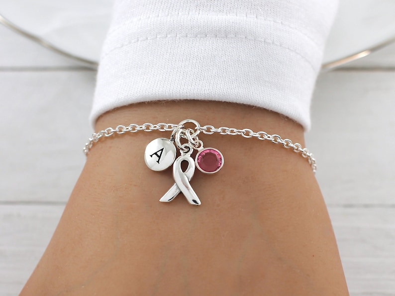 May include: A silver charm bracelet with a pink ribbon charm, a small pink crystal charm, and a silver charm with the letter A.
