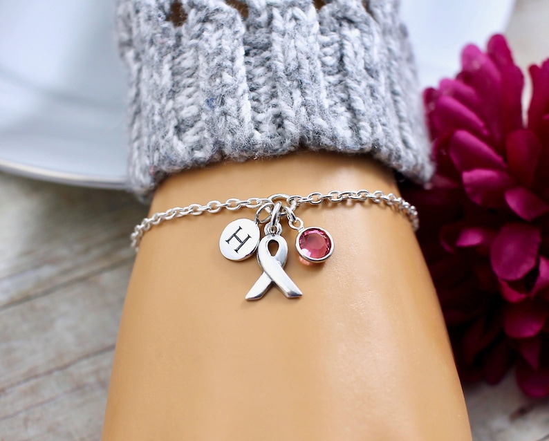 May include: Silver charm bracelet with a pink ribbon, a pink crystal, and a charm with the letter "H".