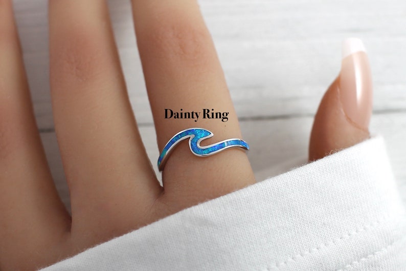 Puede incluir: Un delicado anillo de plata con un dise&ntilde;o de ola de &oacute;palo azul. El anillo est&aacute; en un dedo y el texto "Dainty Ring" est&aacute; por encima del anillo.