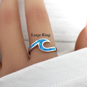 Puede incluir: Un anillo de plata con un dise&ntilde;o de ola de &oacute;palo azul. El anillo est&aacute; en un dedo y el texto "Large Ring" est&aacute; por encima del anillo.