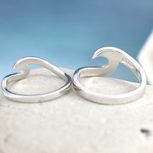 Puede incluir: Dos anillos de plata con forma de ola, uno con la ola hacia arriba y el otro con la ola hacia abajo. Los anillos est&aacute;n sobre una roca blanca.