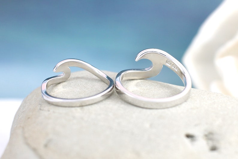 Puede incluir: Dos anillos de plata con forma de ola, uno con la ola hacia arriba y el otro con la ola hacia abajo. Los anillos est&aacute;n sobre una roca blanca.