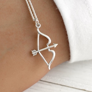 May include: A silver necklace with a bow and arrow pendant. The arrow is pointing to the right and the bow is shaped like a heart.