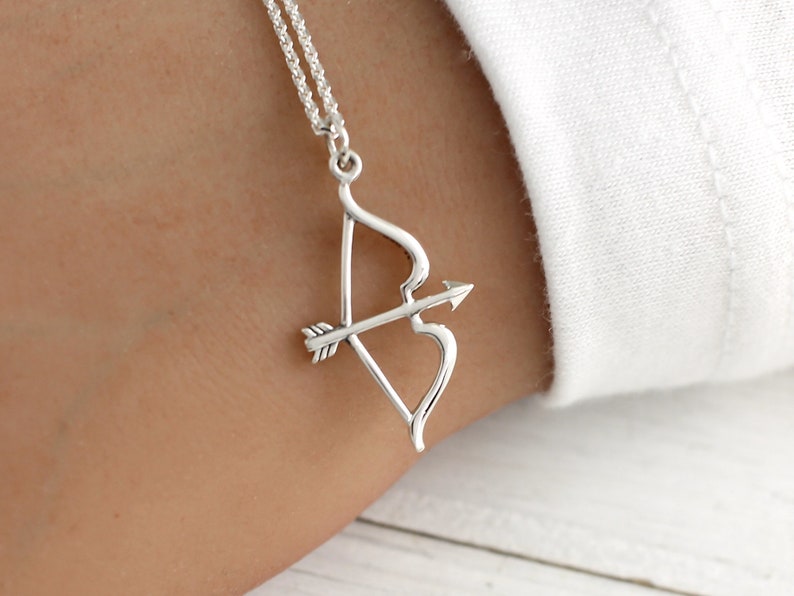 May include: A silver necklace with a bow and arrow pendant. The arrow is pointing to the right and the bow is shaped like a heart.