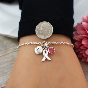 May include: A silver chain bracelet with a pink ribbon charm, a small letter 'L' charm, and a pink crystal charm. The bracelet is shown on a person's wrist.
