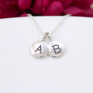 May include: A silver necklace with two circular pendants, each engraved with a single letter, "A" and "B". The pendants are connected by a delicate chain. The background is white with a blurred pink floral accent.