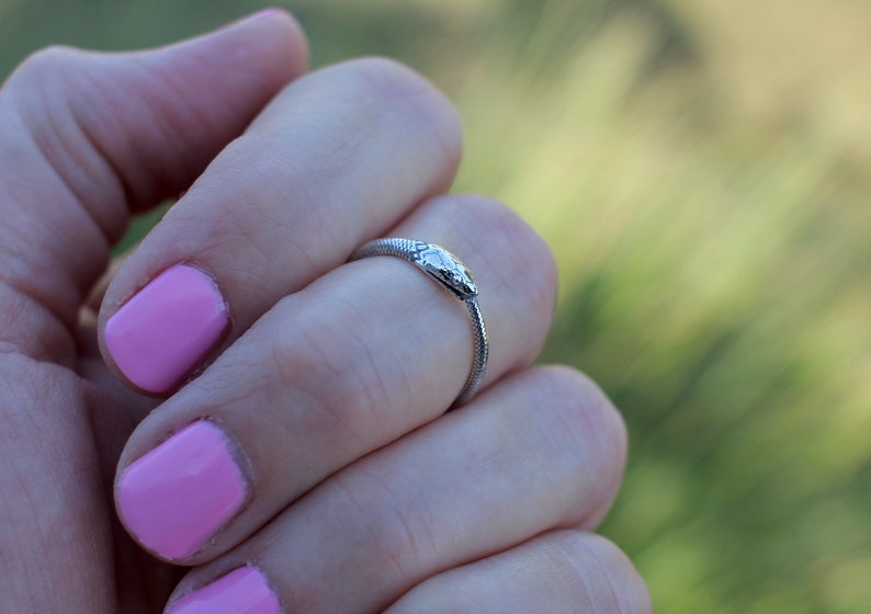 Puede incluir: Un anillo de serpiente de plata con un dise&ntilde;o detallado de cabeza de serpiente. El anillo se lleva en un dedo con esmalte de u&ntilde;as rosa.