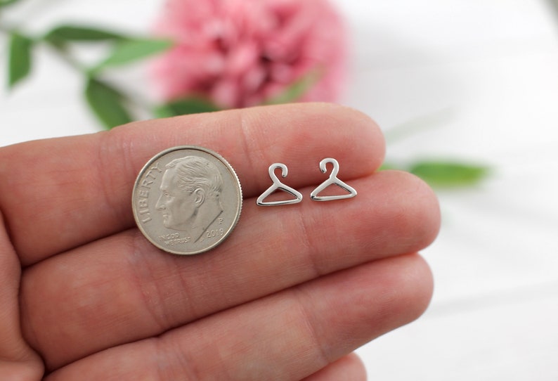 May include: Two silver hanger shaped earrings on a hand with a US dime for scale.