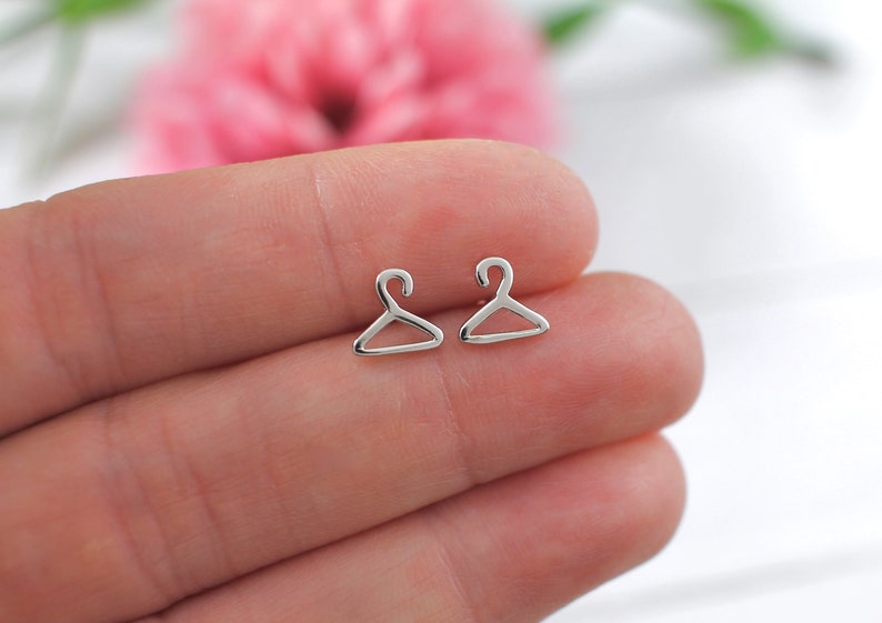 May include: A pair of silver hanger-shaped stud earrings. The earrings are small and delicate, and they are perfect for adding a touch of style to any outfit.