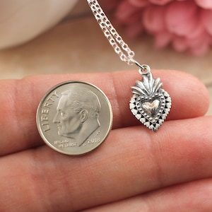May include: A silver necklace with a small, detailed silver heart pendant. The heart has a gold center and is surrounded by a decorative border. The pendant is about the size of a dime.