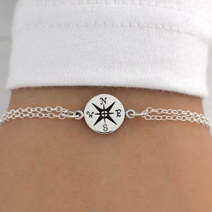 May include: A silver chain bracelet with a compass charm. The compass charm has a black compass rose with the letters N, S, E, and W.