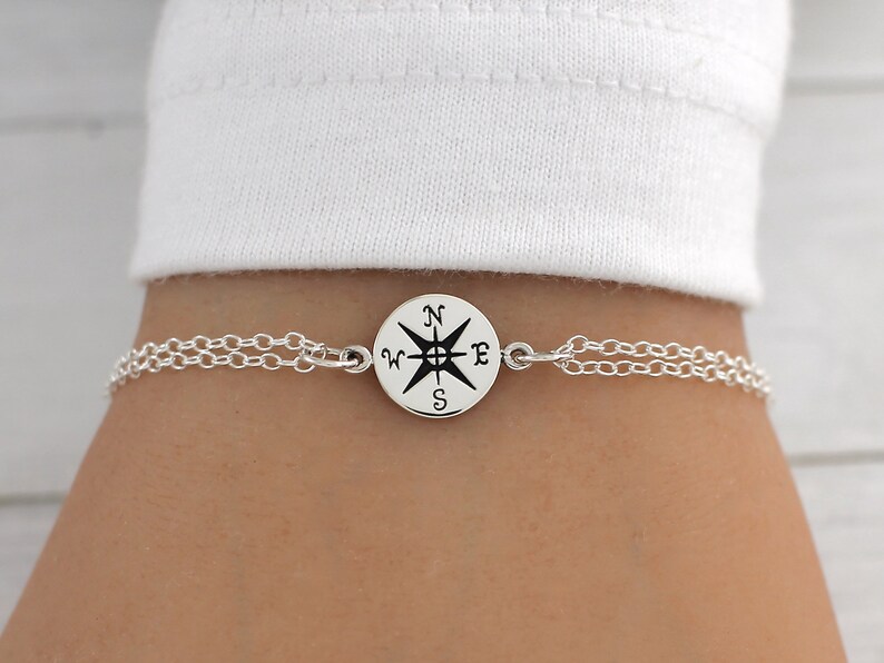 May include: A silver chain bracelet with a compass charm. The compass charm has a black compass rose with the letters N, S, E, and W.