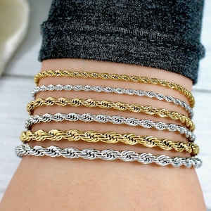 May include: Close-up of several rope chain bracelets in gold and silver tones. The bracelets are of varying thicknesses and are stacked on a wrist. The jewelry is displayed against a neutral background.