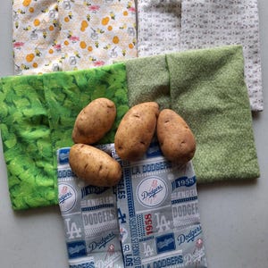 May include: An assortment of fabric potato bags in various patterns, including bees, cats, and Dodgers baseball logos. Several brown potatoes rest on top of the bags, showcasing the product's use.