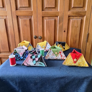 May include: A collection of handmade triangular fabric pouches in various patterns and colors, including floral, geometric, and abstract designs. Each pouch is secured with a button closure. Spools of thread in red and green are also visible.