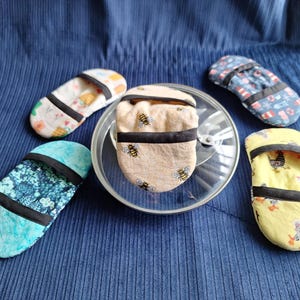 May include: Five colorful oven mitts with various patterns, including bees, chickens, and floral designs, are arranged on a blue surface. Each mitt has a black trim and a loop for hanging. The mitts are designed to fit over a hand.