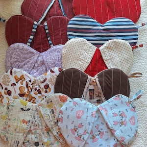 May include: A collection of heart-shaped quilted pot holders in various patterns and colors. Designs include stripes, floral, and novelty prints. Each pot holder has a loop for hanging.