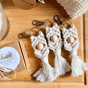 May include: A collection of handmade items, including three macrame keychains with wooden beads and tassels, a jar of body butter, a loofah, and a small box with a sticker. The items are arranged on a wooden surface.