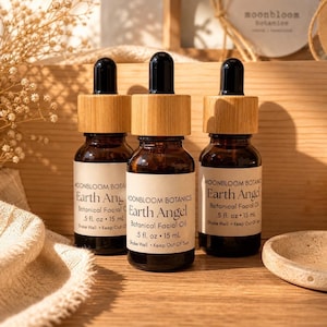 May include: Three small brown glass bottles with bamboo lids and black droppers. Each bottle has a white label with the text "Earth Angel Botanical Facial Oil". The bottles are displayed on a wooden surface.