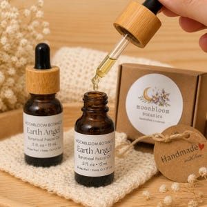 May include: Two small brown glass bottles of Earth Angel Botanical Facial Oil with wooden caps. A dropper dispenses the oil into one bottle. The label reads "Moonbloom Botanics". A small box and a tag that says "Handmade with love" are also visible.